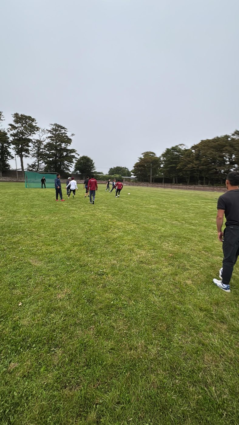 Ishchilar lager chekkasidagi oʻtli maydonda futbol oʻynashadi; uzoq tomondagi darvozalar, maydon ortida esa daraxtlar qatori bor