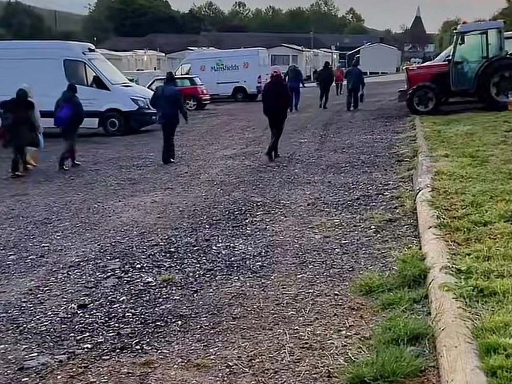 Seasonal workers walking to their shift across the gravel yard of Mansfield farm: minibuses (one with Mansfields logo), red tractor, with residential caravans and a packhouse in the background